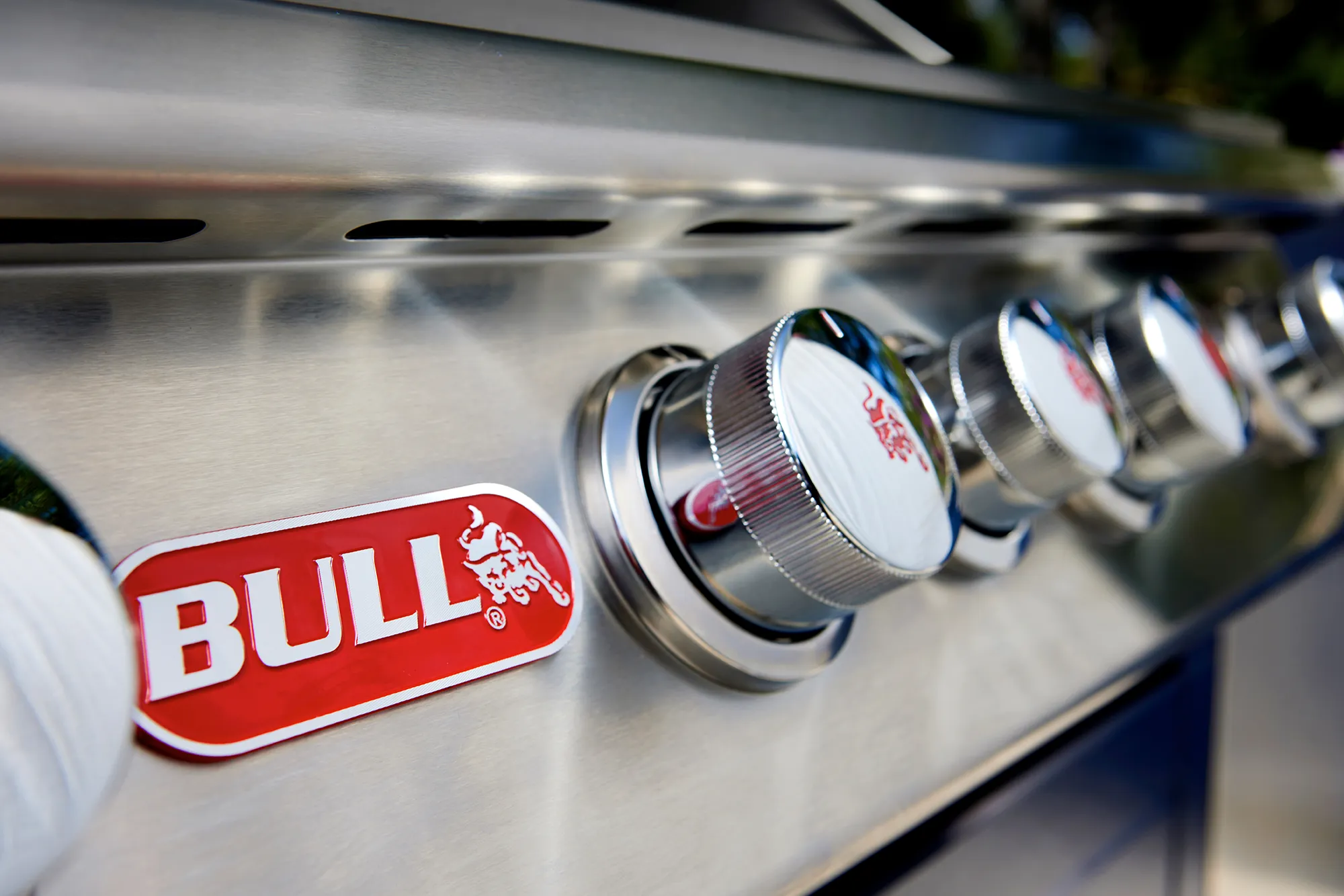 Close-up of the red Bull BBQ badge and polished chrome control knobs with embossed bull emblems on a 304 stainless steel grill control panel, outdoor patio background.
