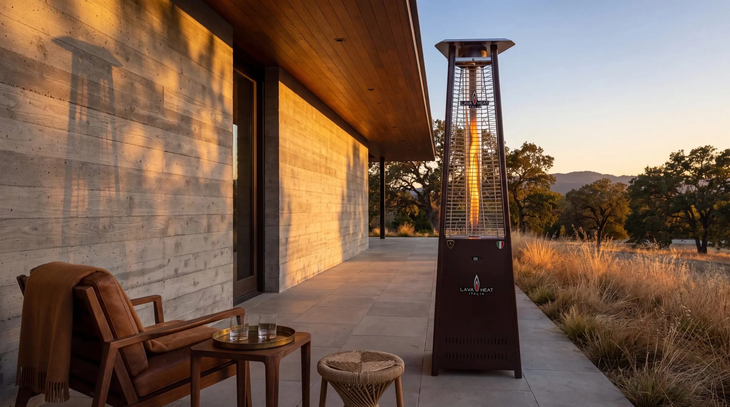 Lava Heat Italia 2G flame-tower patio heater on a Napa-style patio at sunset — glass column with visible column of flame, concrete and timber architecture, mid-century chair with throw blanket.