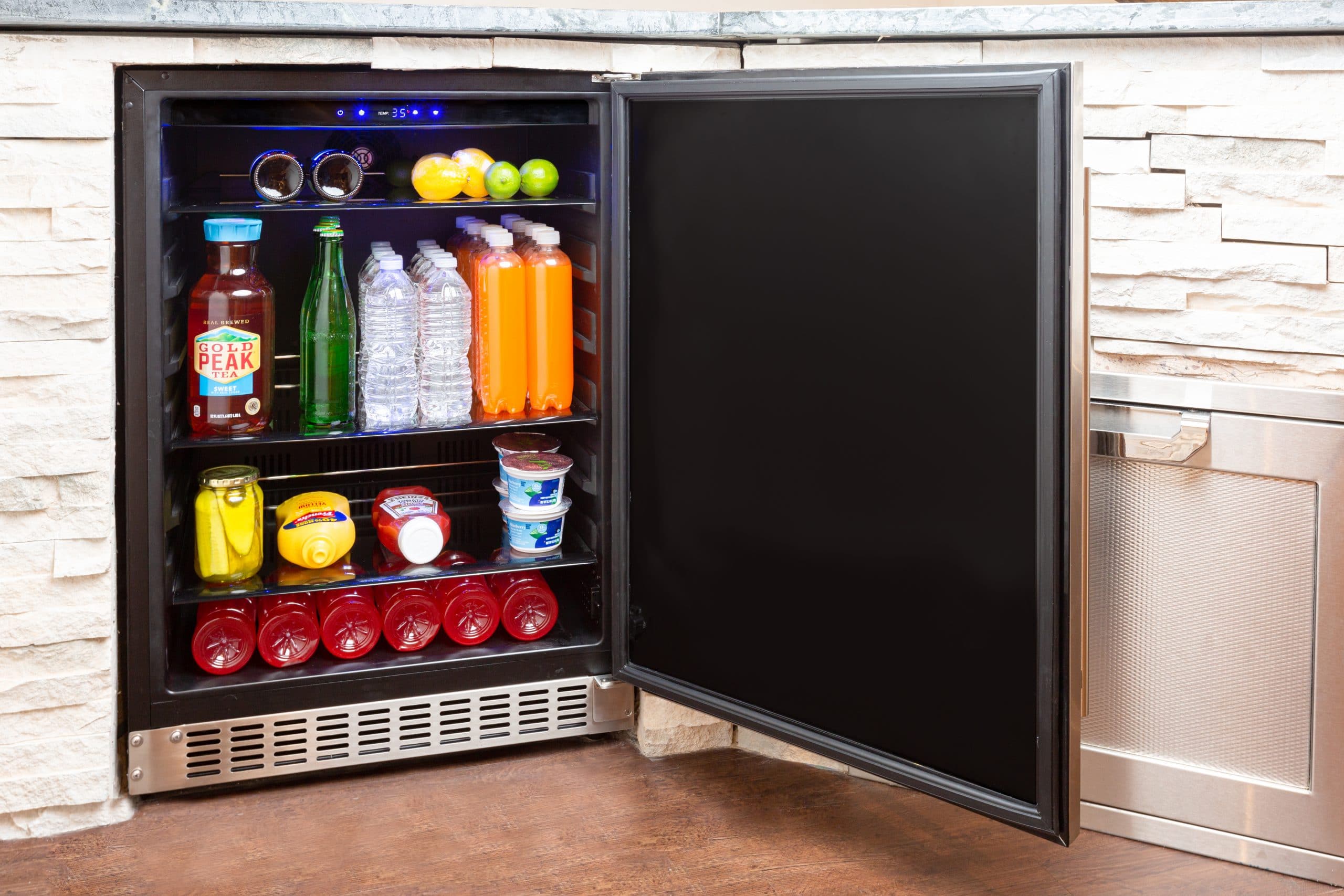 Azure dual-zone beverage and wine center integrated into a stone outdoor kitchen counter, glass French doors showing rows of chilled bottles — poolside evening scene.