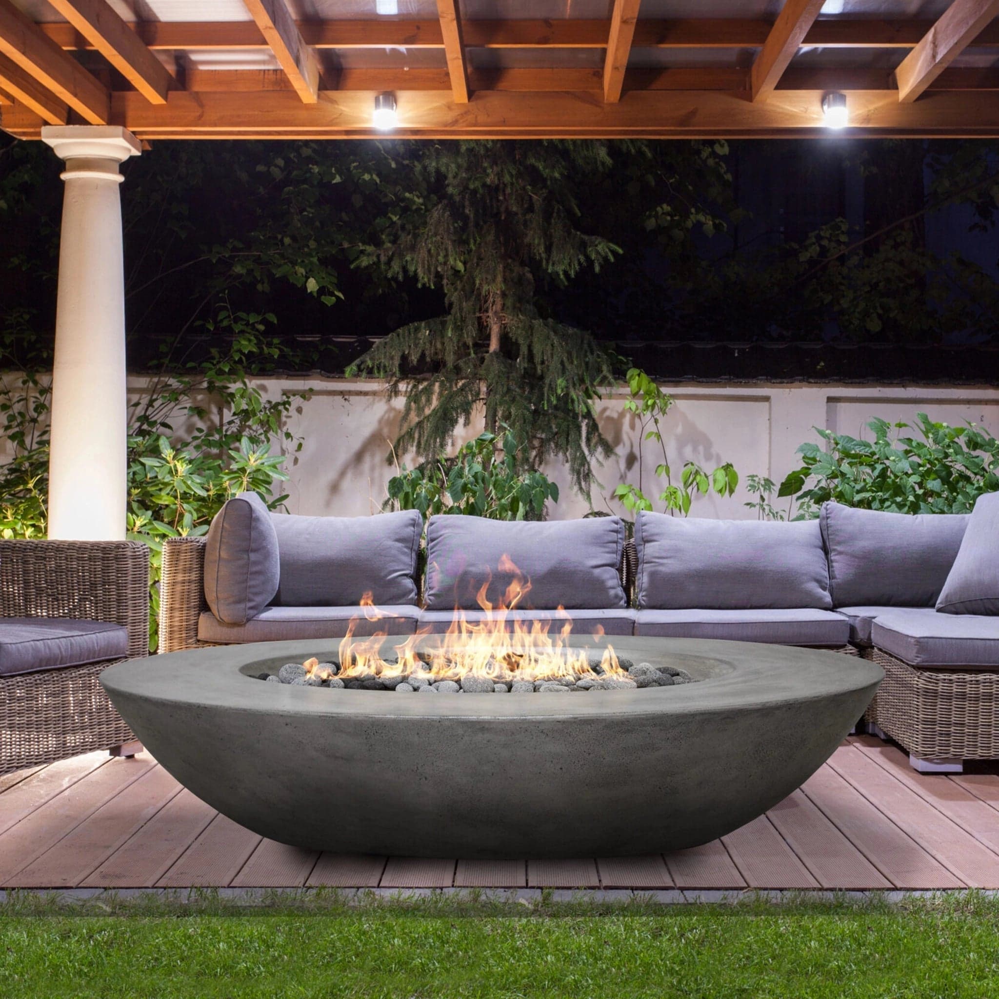 Prism Hardscapes Ovale GFRC concrete gas fire bowl burning under a wooden pergola with cushioned sectional seating — the sealed-base gas pit this article recommends for covered patio installs.